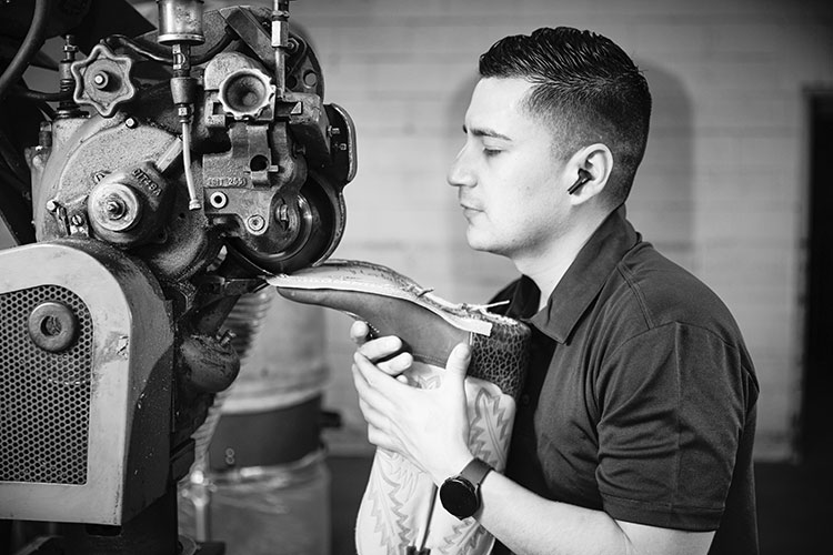 A black-and-white image of a young Hispanic man holding a boot as he’s working on building it.